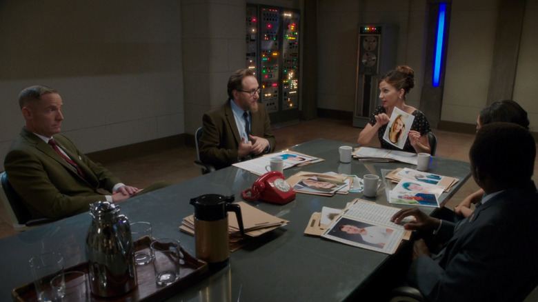 Val holds up a headshot of a Bad Place resident while sitting at a board room with demon boss Shawn and her coworkers Glenn, Babadjan and Gayle in 