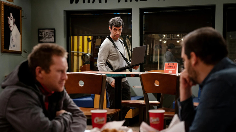 Nathan Fielder wears a laptop strapped to his body while observing two people talking over lunch in the foreground in 