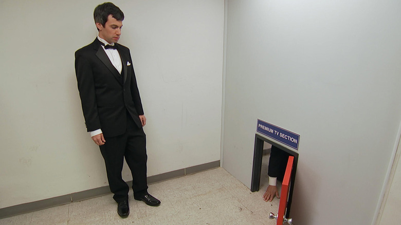 Nathan Fielder wears a tuxedo while standing next to a man kneeling on the other side of a miniature door labeled Premium TV Section in 
